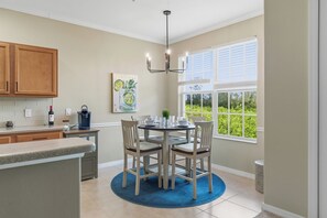 A kitchen with wooden cabinets, a dining table with chairs, a blue rug, and a window with a view of greenery. - Majestic Palms-Close to Beaches, Heated Pool & Spa, Fully Updated Interior (Fort Myers)