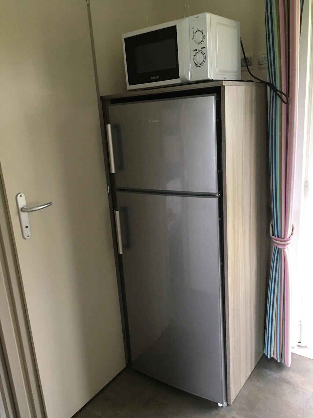 A compact refrigerator with a microwave on top, a door handle, and a striped curtain in the background.