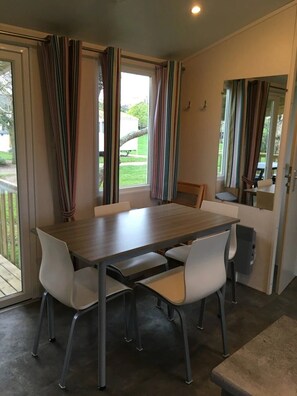 Ein Essbereich mit einem Holztisch, vier weißen Stühlen, großen Fenstern mit gestreifter Gardine und Blick auf einen Garten.