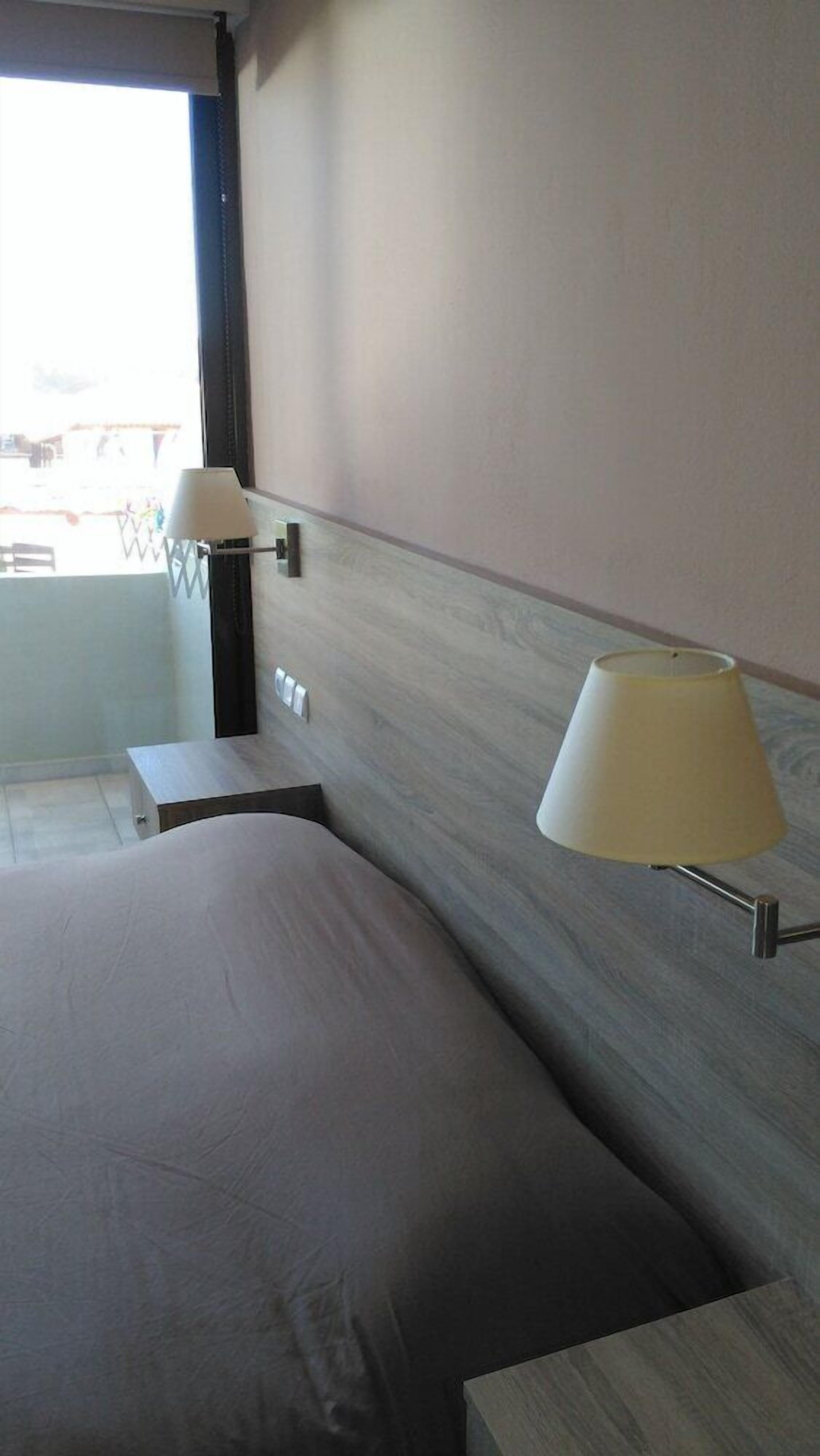 A hotel room with a bed, a nightstand, a lamp, and a view of the outside.