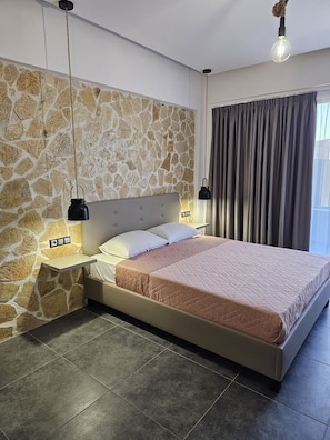 A bedroom with a stone wall, a bed with a beige headboard, two bedside tables, and a large window with curtains. - Santo George Beach (Malevizi)