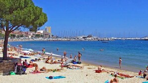 Una playa con gente tomando el sol y nadando, un muelle con barcos, y edificios al fondo.