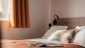 Un dormitorio con cama, almohadas, mesita de noche, lámpara y ventana con cortinas.