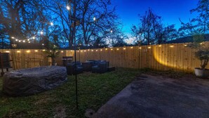 Un jardin avec une clôture en bois, des guirlandes lumineuses, du mobilier d’extérieur et un barbecue abrité.