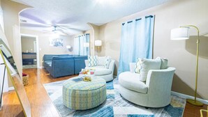 Un salon avec un canapé bleu, un fauteuil blanc et un pouf à motifs. On y trouve un ventilateur de plafond, une lampe sur pied et un tapis.