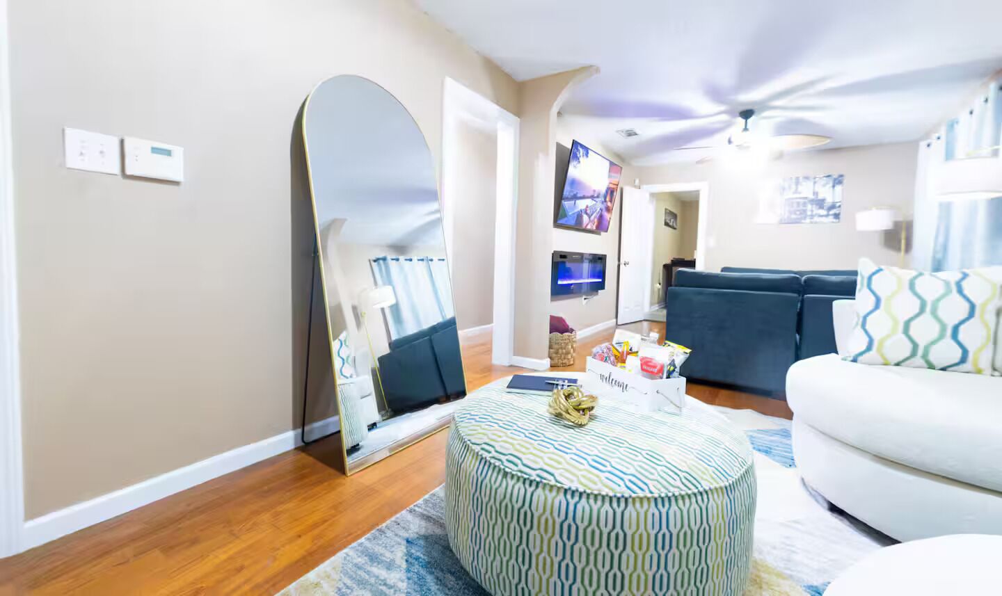 Un salon avec un pouf rond, un canapé blanc, un grand miroir et un téléviseur.