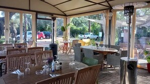 Área de pátio ao ar livre com mesas e cadeiras de madeira, amplas janelas e vista para carros estacionados e vegetação.