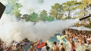 Uma área de piscina ao ar livre movimentada, com pessoas aproveitando a água e um efeito de fumaça ao fundo.