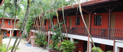 A row of red buildings with balconies, surrounded by palm trees and gravel paths.