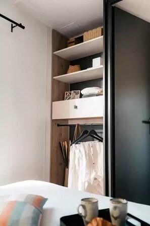 Ein Schlafzimmer mit einem Bett, einem Nachttisch mit Tasse und Buch, einem Schrank mit Regalen und Kleiderhaken sowie einer Tür.