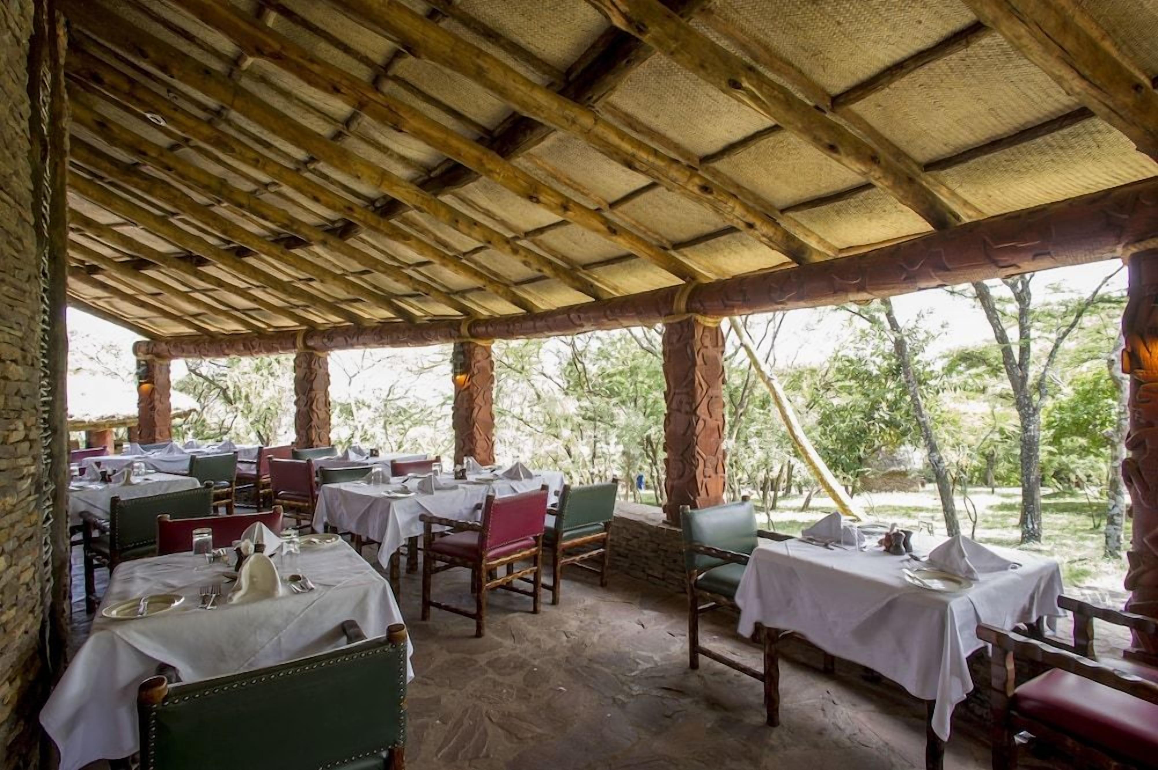 Un espacio de comedor al aire libre con mesas y sillas, cubierto por un techo de madera, rodeado de follaje.