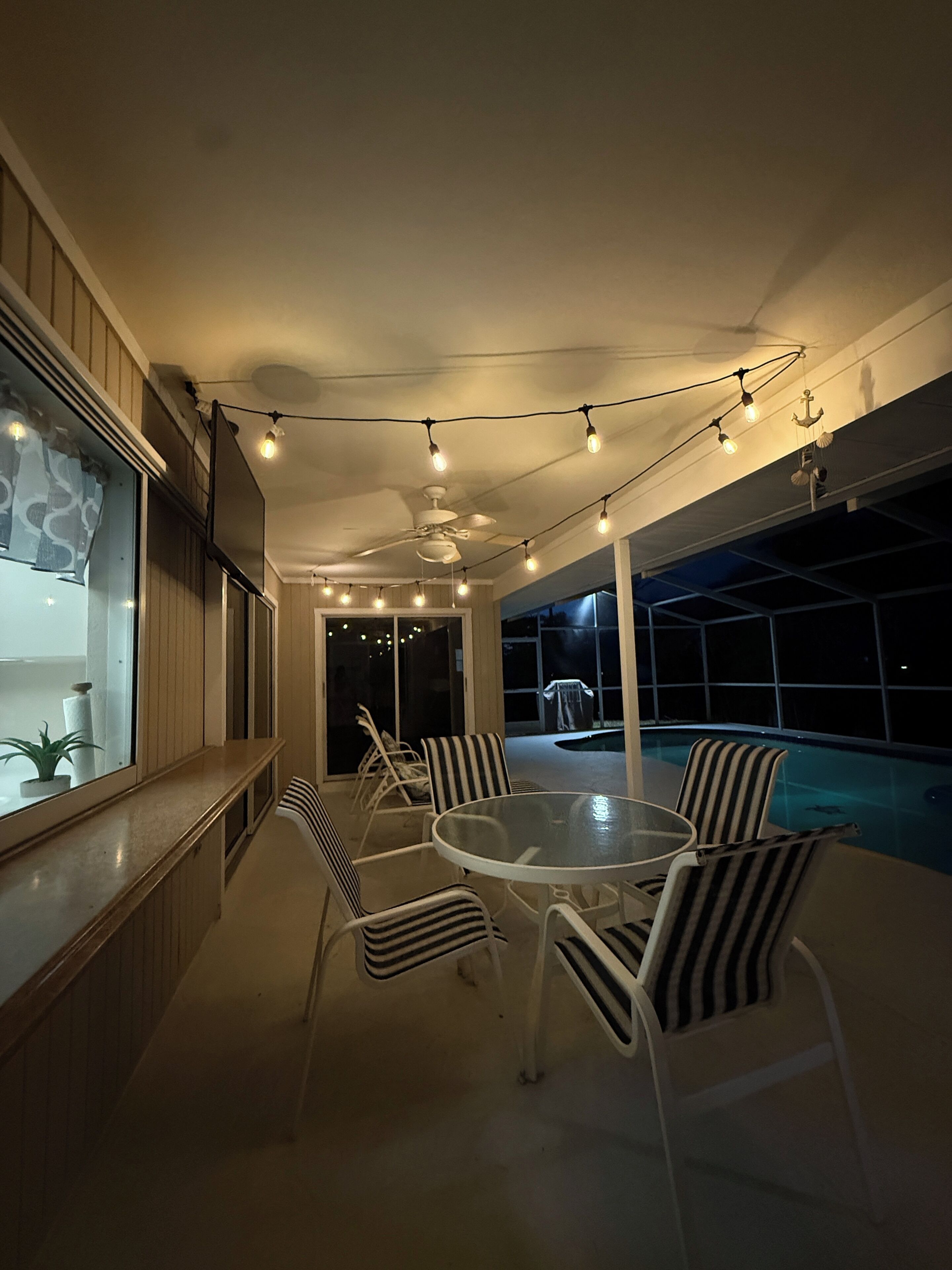 Un espace aménagé au bord de la piscine, avec une table ronde en verre et des chaises à rayures, des guirlandes lumineuses et un ventilateur de plafond.