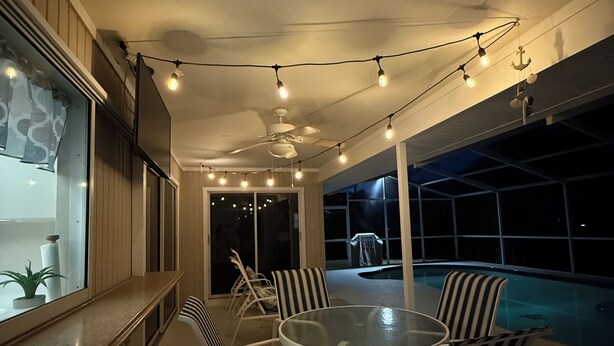 A poolside area with a round glass table and striped chairs, string lights, and a ceiling fan.
