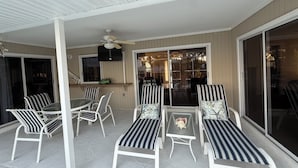 A covered patio area with striped lounge chairs and a table, a ceiling fan, and a TV mounted on the wall.