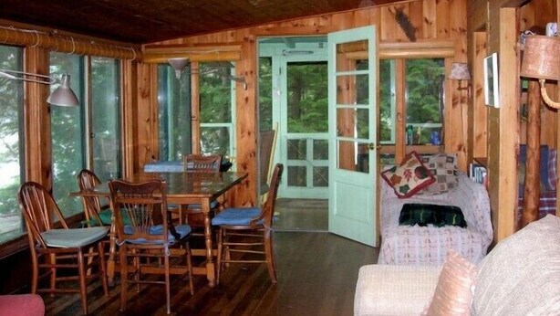 A wooden cabin interior with a dining area, a sofa, and a door leading outside. - Shelving Rock Camp on Squam Lake (Center Harbor)