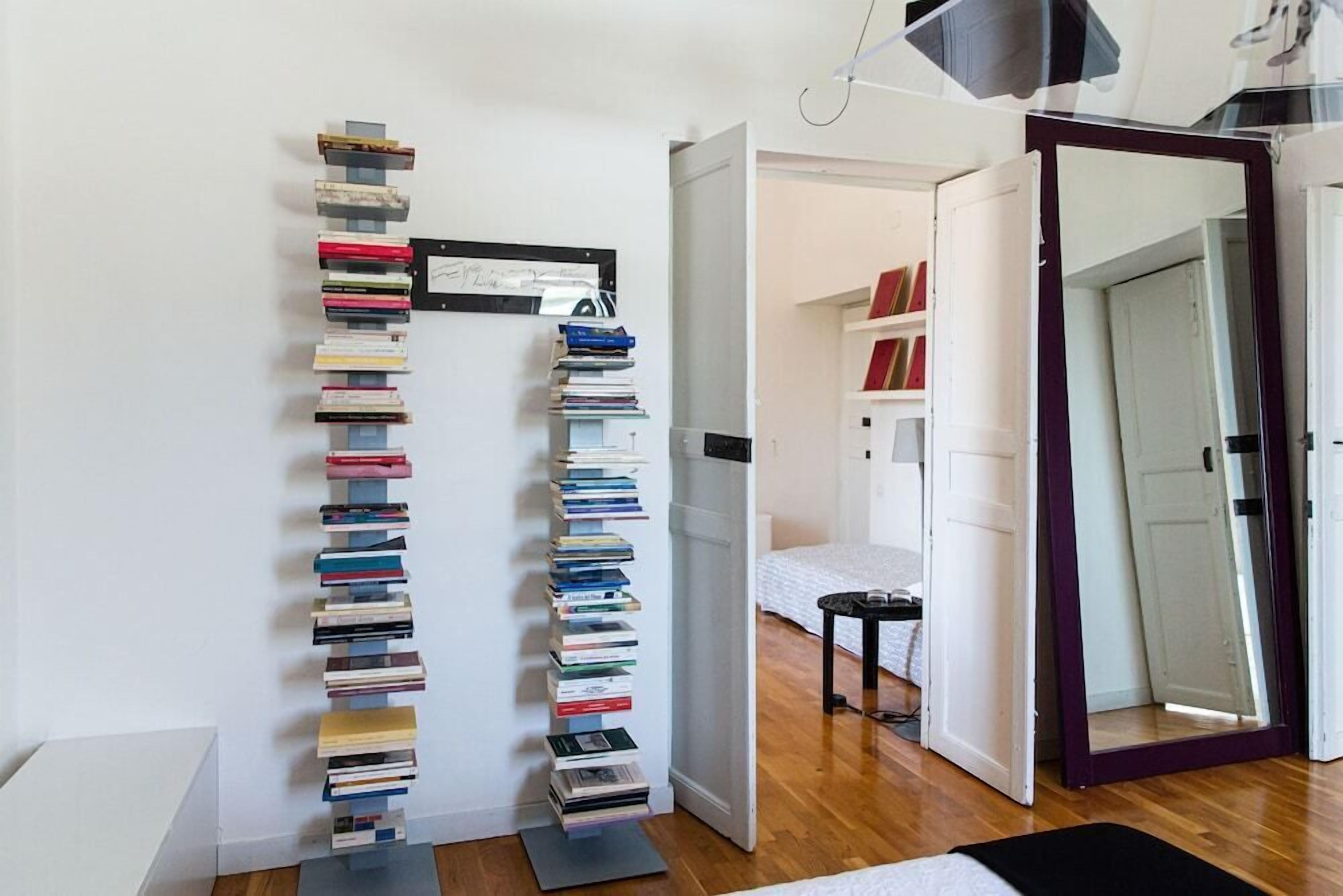 Una habitación con estantería, cama, una mesita y un espejo.
