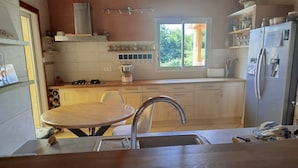 Et køkken med spisebord, vask, komfur, køleskab og et vindue med udsigt til grønne omgivelser.