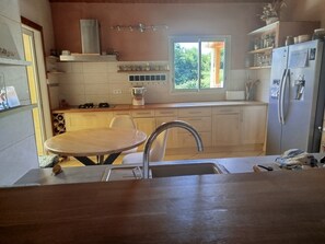 A kitchen with a dining table, a sink, a stove, a refrigerator, and a window with a view of greenery. - Charming Nest for 2 in the Heart of Lodève with Mountain View, Shared Pool & Wi-Fi (Lodève)