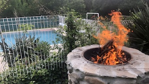 En pejsesten med flammer, omgivet af et metalhegn, nær en swimmingpool.