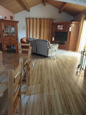 Una sala de estar con techo de madera, un sofá, un televisor y un comedor con sillas y un armario.