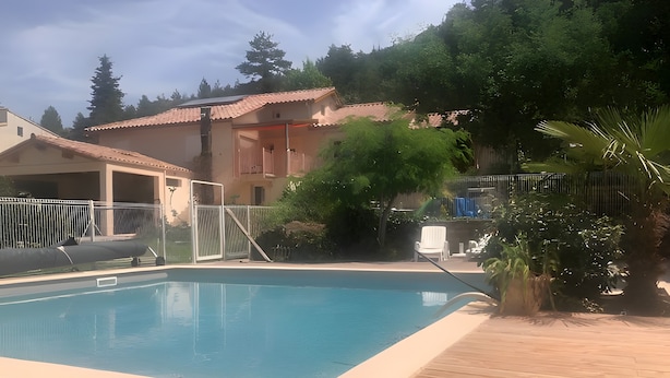 En swimmingpool med træterrasse, omgivet af et hegn, og en bygning med et tegltag i baggrunden.