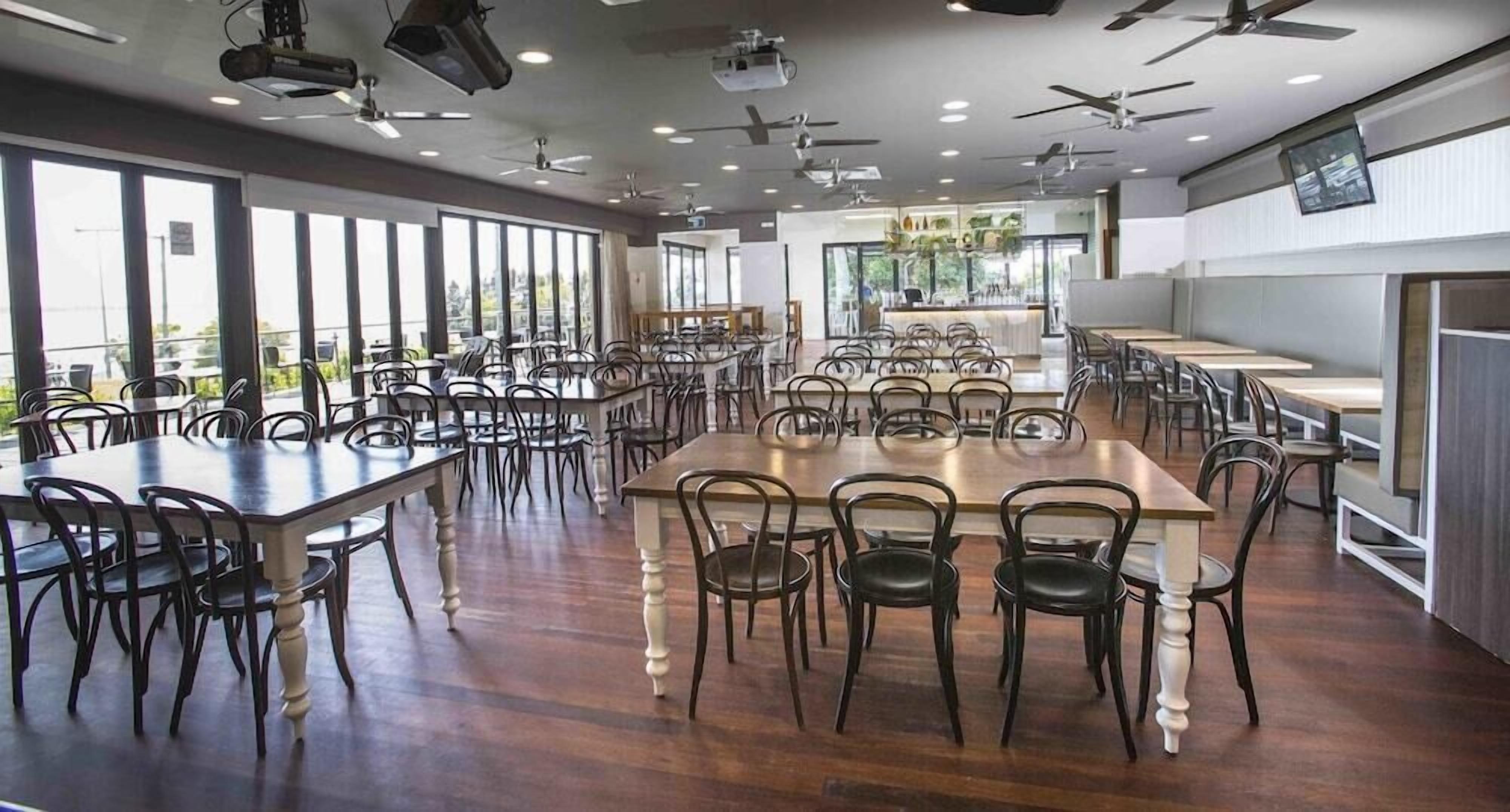 A spacious dining area with multiple tables and chairs, ceiling fans, and large windows providing natural light.
