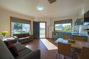 A modern living room with a sofa, dining table, and chairs, a kitchen with a window, and a door leading outside. - West Beach Parks Resort (Adelaide)