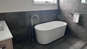 A modern bathroom with a freestanding bathtub, a window above the sink, and dark tiled walls. - West Beach Parks Resort (Adelaide)