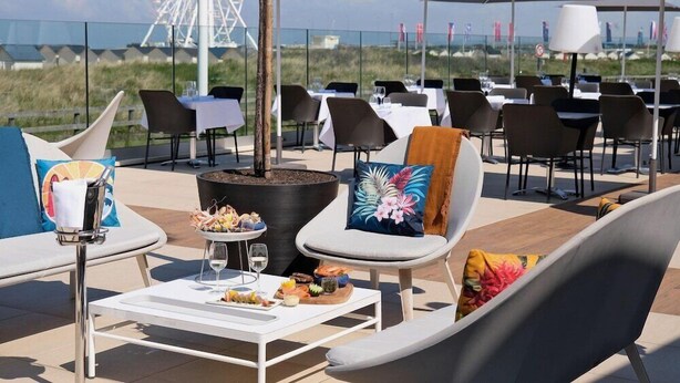 Un espace extérieur aménagé pour les repas, avec des tables et des chaises, un foyer et une vue sur la plage, ainsi qu’une grande roue en arrière-plan.