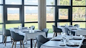 Un restaurant doté de grandes fenêtres donnant sur un vignoble et des champs. Les tables sont dressées avec des nappes blanches et des chaises noires.