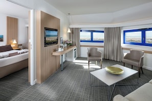 Une chambre d’hôtel équipée d’un lit, d’une télévision, d’une table basse, de deux fauteuils, d’un bureau et de grandes fenêtres offrant une vue dégagée.