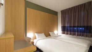 Een hotelkamer met een houten hoofdbord, een bed met witte lakens, een nachtkastje en een raam met gestreepte gordijnen.