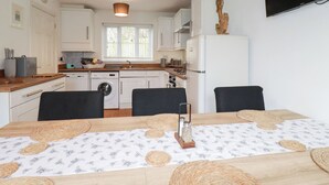 A compact kitchen with white cabinets, a built-in oven, and a refrigerator. There is a dining table with a white tablecloth and woven placemats. - Poldam (St Austell)