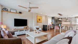 Een moderne woonkamer met een flatscreen televisie, een plafondventilator, een eethoek met een tafel en stoelen, en een keuken op de achtergrond.