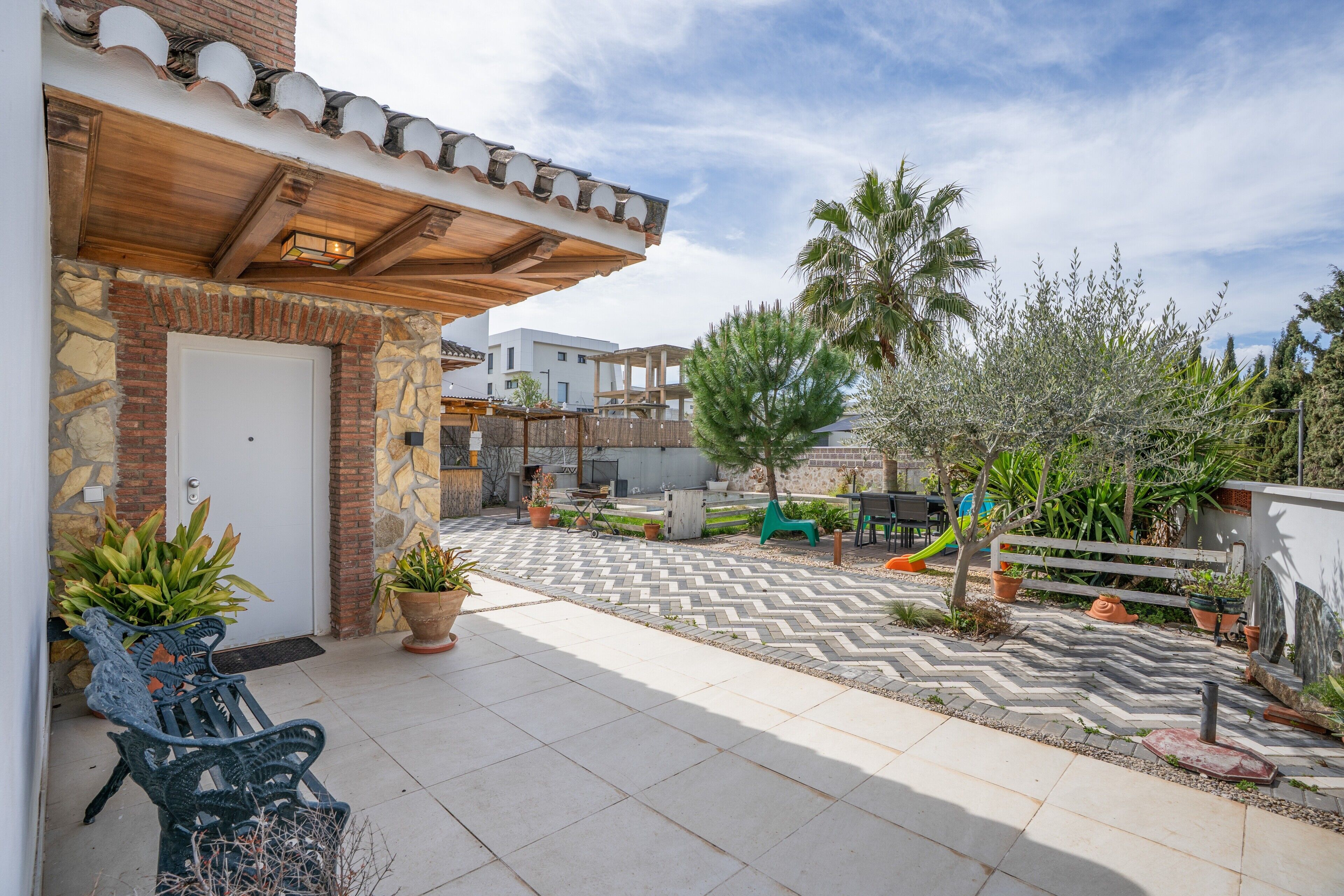 Un patio con suelo de baldosas, una pérgola de madera, un banco y plantas en macetas.