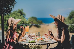 Deux personnes sont assises à une table en plein air, devant une variété de plats et de boissons, et elles jouissent d’une vue panoramique sur un paysage magnifique.