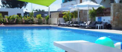 En swimmingpool med liggestole og parasoller, omgivet af en bygning med en stenfassade.