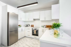 A modern kitchen with white cabinets, a stainless steel refrigerator, a built-in oven, and a countertop with a plant and glasses. - Latchi Escape Hotel and Suites (Neo Chorio)