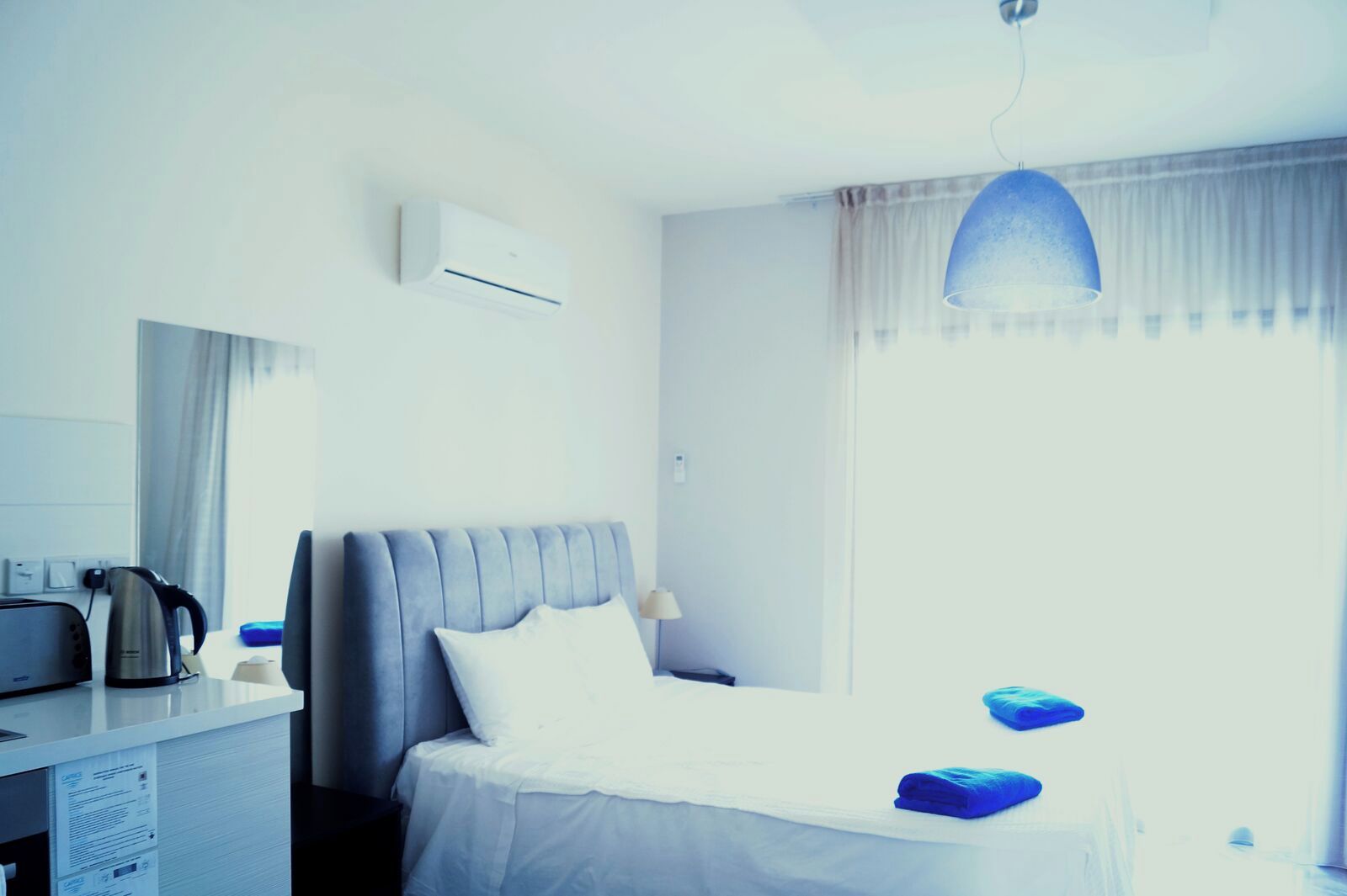 Una habitación de hotel con una cama, un escritorio con un hervidor, una lámpara y una ventana con cortinas.
