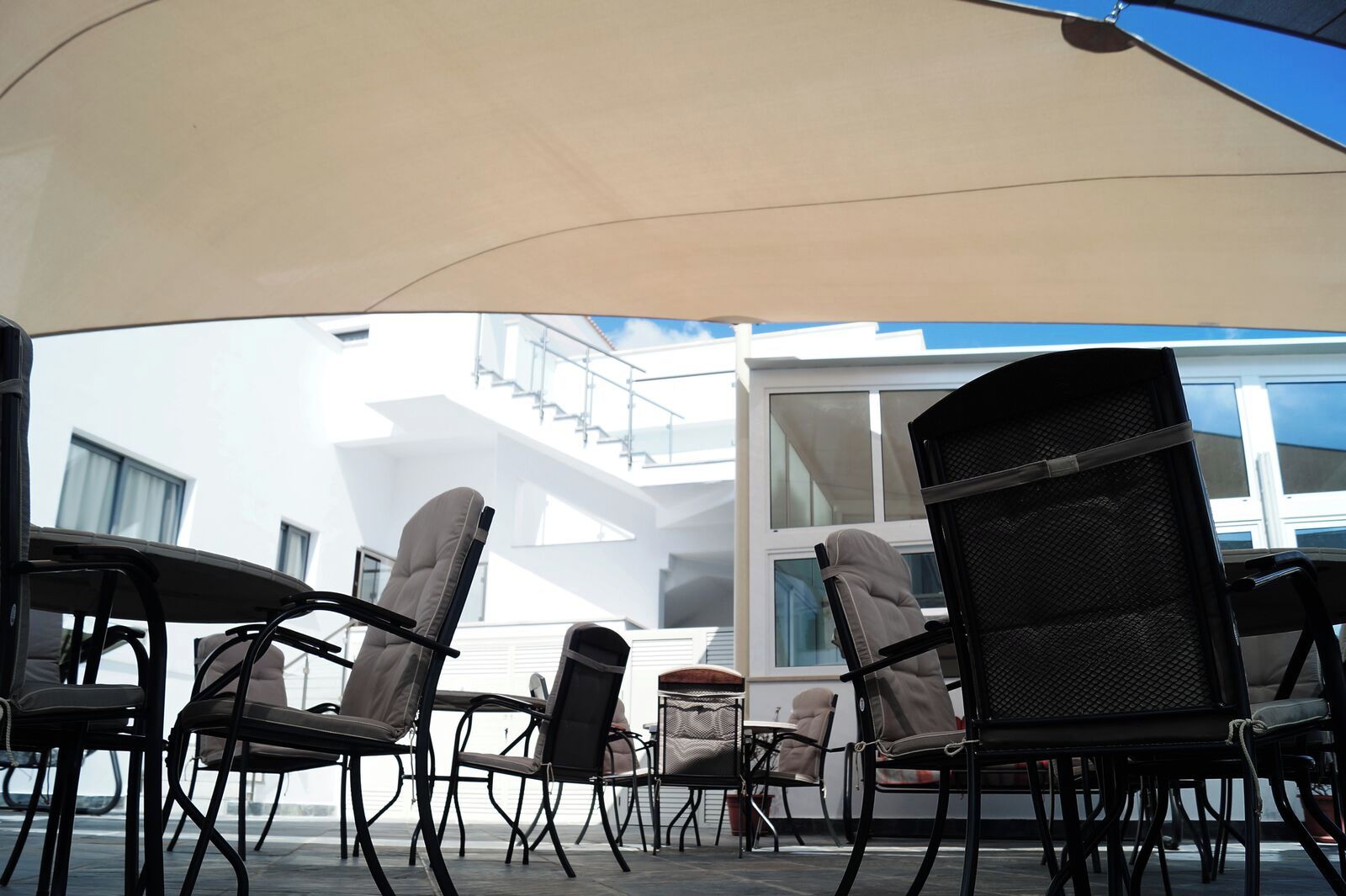 Mobiliario exterior con sillas y mesas bajo un toldo, con vistas a un edificio con ventanas amplias y un balcón.
