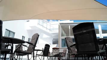 Mobiliario exterior con sillas y mesas bajo un toldo, con vistas a un edificio con ventanas amplias y un balcón.