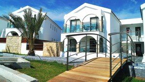 A modern two-story building with a wooden deck, glass balustrades, and a stone wall. - Latchi Escape Hotel and Suites (Neo Chorio)
