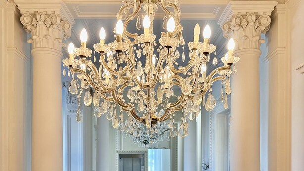 A grand hallway with a large chandelier, ornate columns, and a patterned floor leading to a room with a chair and a table. - Hotel Universal (Livorno)