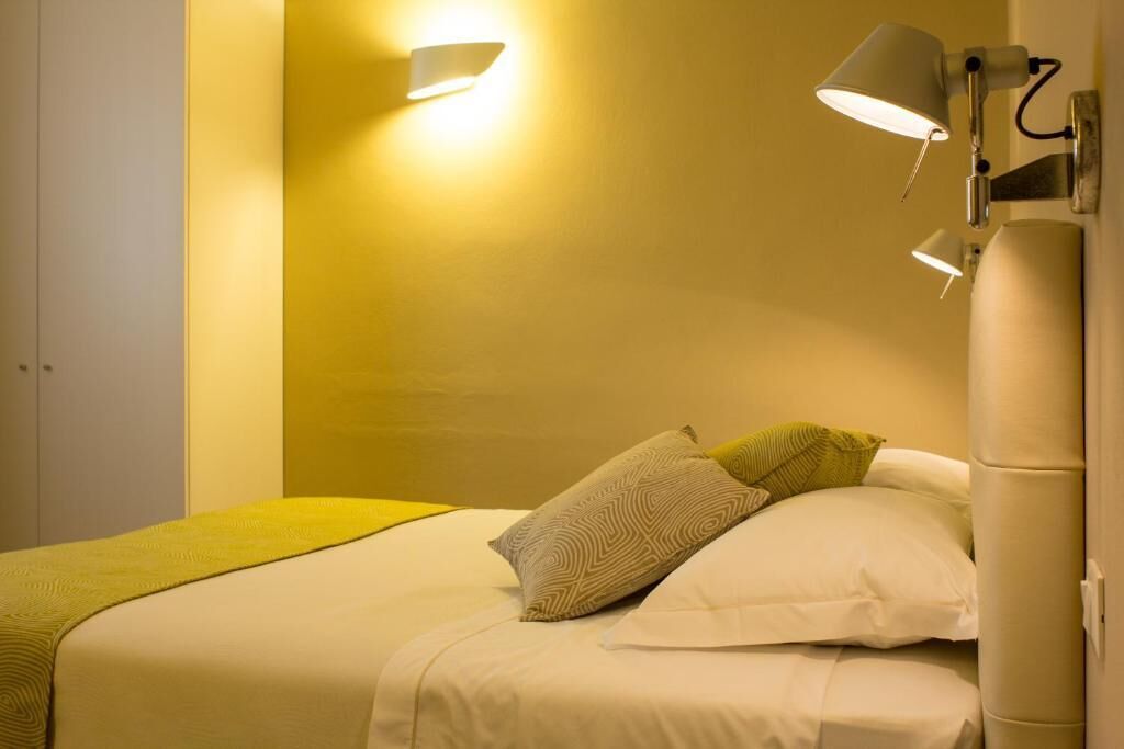 A neatly made bed with a yellow bedspread and two pillows, a wall-mounted lamp, and a bedside table with a lamp.