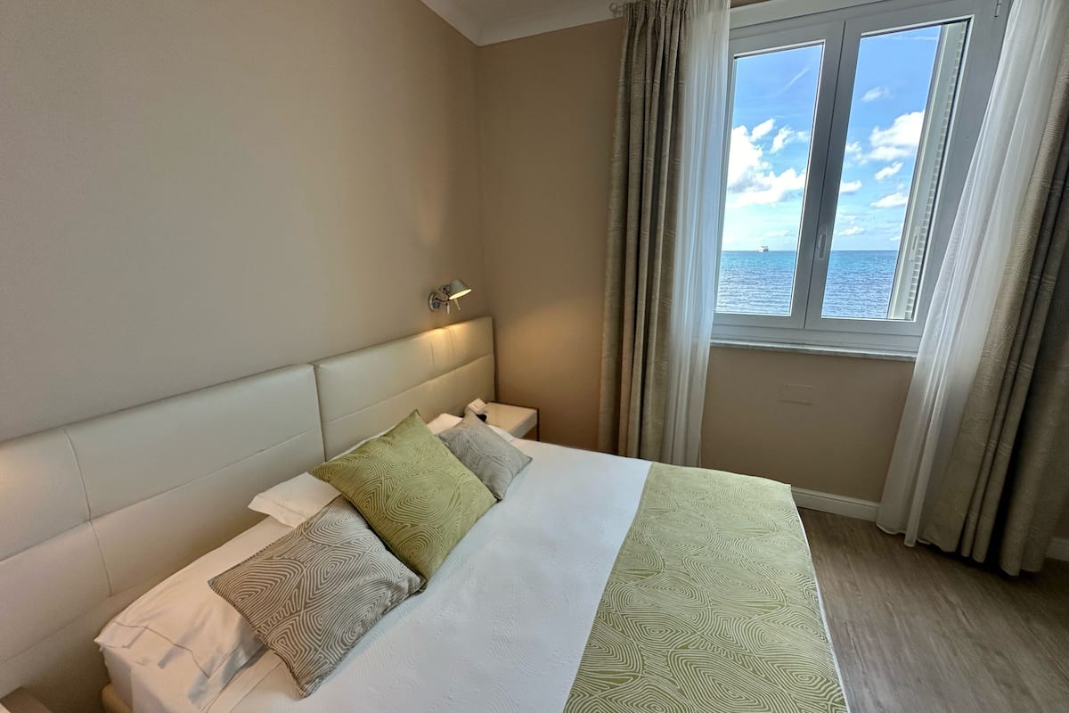 Una habitación de hotel con cama, mesita de noche, lámpara y ventana con cortinas.