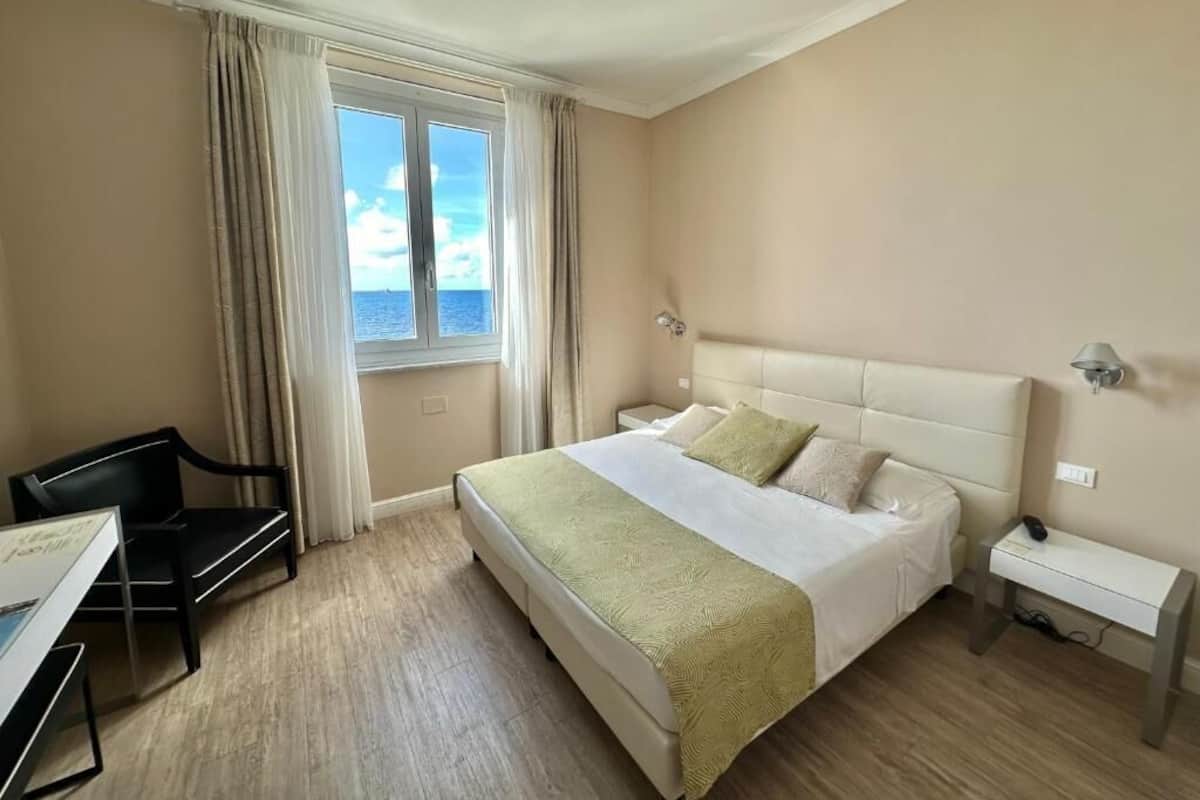 Una habitación de hotel con cama, escritorio, silla, ventana con cortinas y vista al mar.