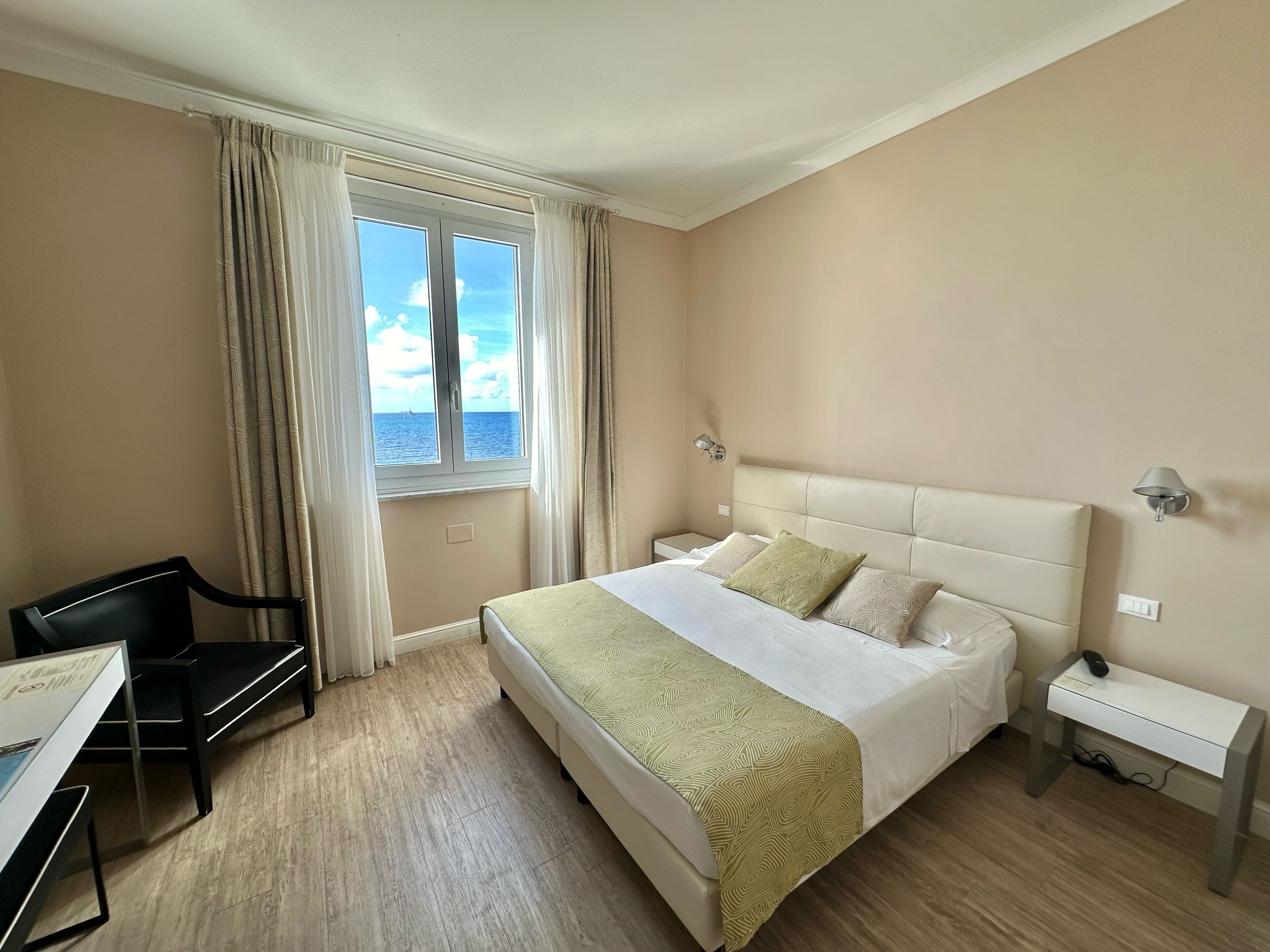 A hotel room with a bed, a desk, a chair, a nightstand, and a window with curtains.