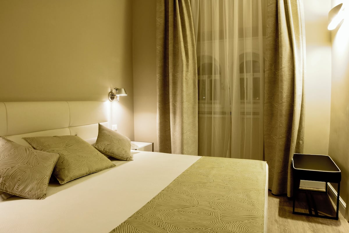 Habitación de hotel con cama, almohadas, mesita de noche, lámpara y ventana con cortinas.