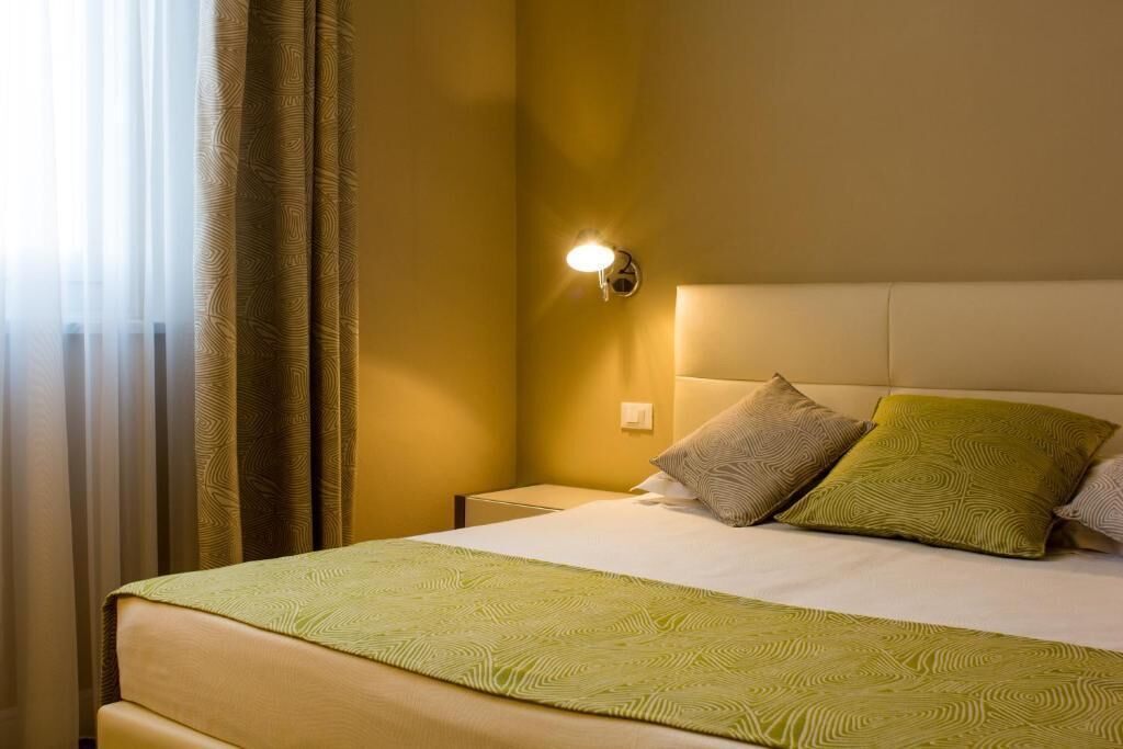 Habitación de hotel con cama, mesita de noche, lámpara de pared y ventana con cortinas.