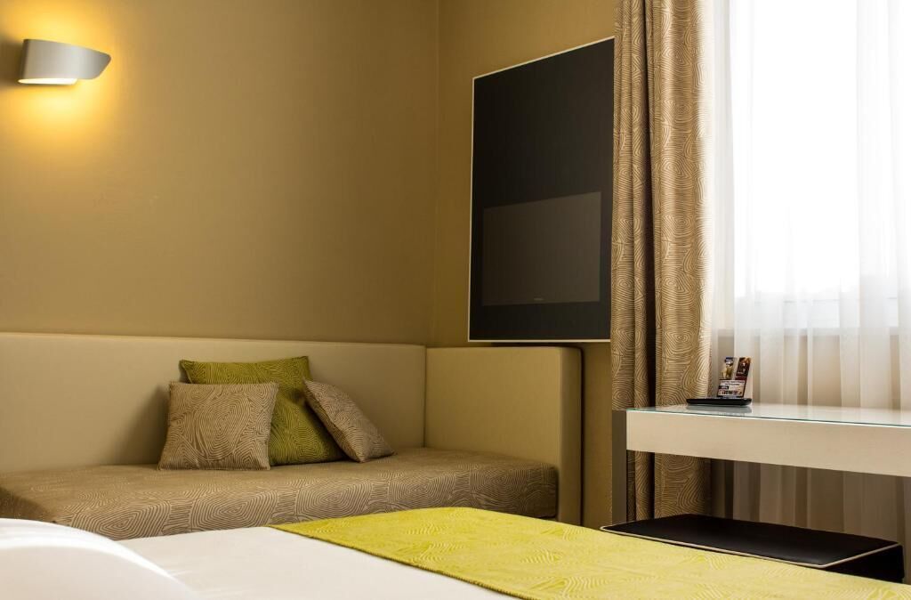 Ein Hotelzimmer mit Sofa, Flachbildfernseher an der Wand, Nachttisch und einem Fenster mit Vorhängen.